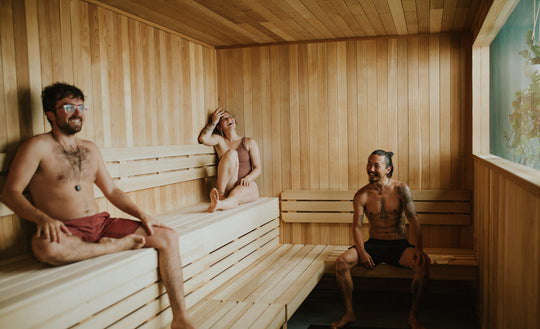 Three friends sitting in a natural wood sauna and laughing together. Light it pouring int through a window and plants are visible on the other side.