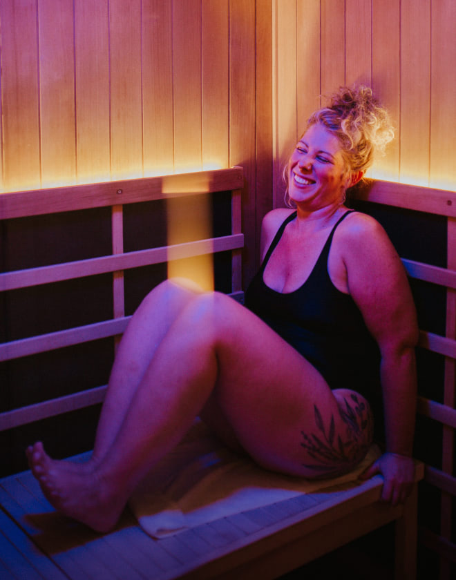 Person sitting in a private infrared sauna cabin with red and purple light, smiling