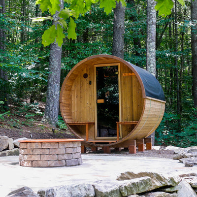 Barrel sauna with a porch on a patio with a fire pit in front of it in a woodsy area